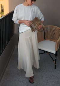 Lys grå oversized t-shirt, beige lang nederdel og brun crossbody taske med kæde. Sorte flip-flops. Vævet stol i baggrunden.