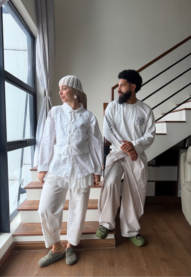Deux personnes en tenue blanche se tiennent près d'un escalier et d'une grande fenêtre, la femme regarde dehors tandis que l'homme la regarde.