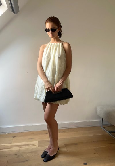 Vestido blanco sin mangas con bordados florales, clutch negro y zapatos planos negros de punta cuadrada, sobre un fondo neutro con suelo de madera.