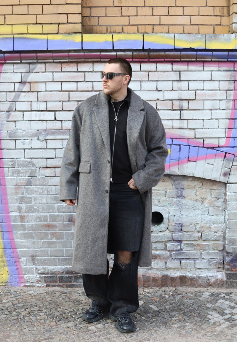 Hombre con un abrigo gris grande, vestimenta negra y gafas de sol, de pie contra una pared de ladrillos cubierta de grafitis en una calle empedrada.