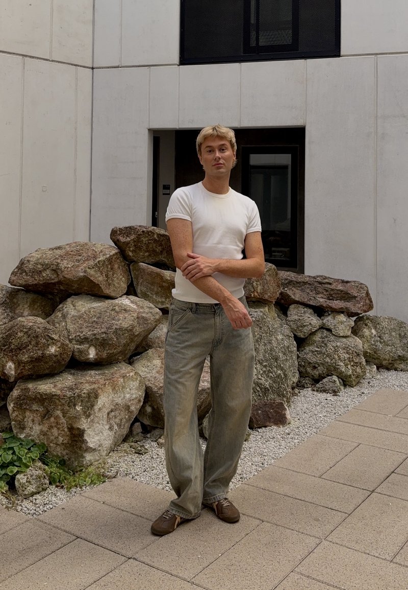 Ung man i vit t-shirt och lösa jeans står med korslagda armar nära stora stenar utanför en modern byggnad.