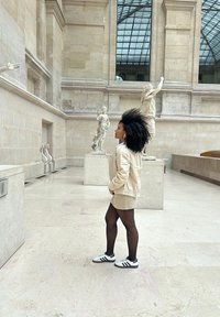 Une personne se tient dans un musée, portant une veste crème et une mini-jupe, avec des collants noirs et des baskets blanches à rayures noires. Des statues l'entourent.