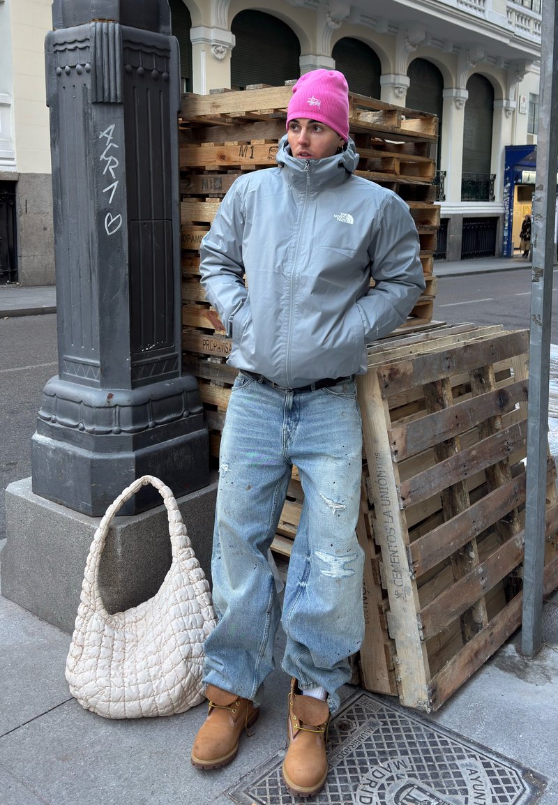 Personne portant une veste grise, un bonnet rose, un jean déchiré et des bottes marron, appuyée sur des palettes en bois sur un trottoir en ville près d'un poteau en pierre et d'un sac blanc.