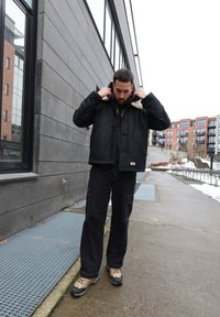Homme ajustant le col d'une veste matelassée noire debout sur un trottoir urbain près d'immeubles modernes, avec des plaques de neige à proximité.