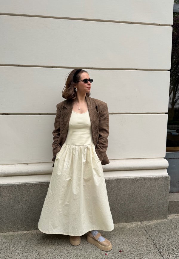 Olkapäille ulottuvat hiukset omaava nainen, jolla on aurinkolasit, kermanvärinen mekko, ruskea ruutukuosinen bleiseri ja beiget korolliset kengät, seisoo valkoista seinää vasten.