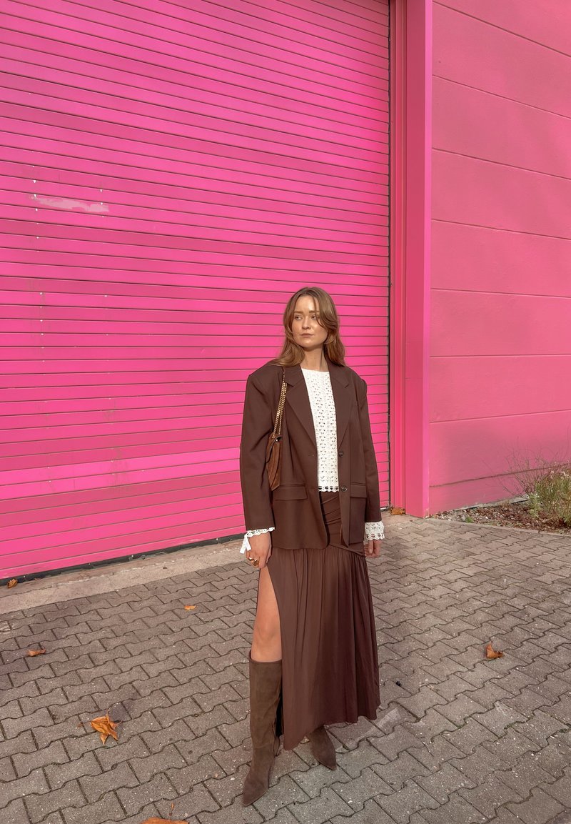 Blazer marron sur une blouse blanche en dentelle, associée à une jupe marron avec une fente latérale. Bottes marron montant jusqu'aux genoux complètent le look sur un fond rose.