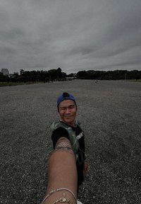 Osoba w czarnej koszuli i zielonej kamizelce, nosząca niebieską czapkę, stoi na żwirowym terenie z drzewami w tle pod pochmurnym niebem.