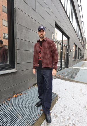 Camisa de franela a cuadros rojos y negros sobre una camiseta negra con diseño gráfico, combinada con vaqueros azul oscuro y zapatos negros, con una pared gris de fondo.