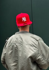 Gorra snapback roja con el logo blanco de "Nueva York", llevada con una chaqueta bomber gris, vista desde atrás contra un fondo verde oscuro.