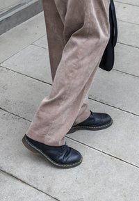 Μαύρα δερμάτινα παπούτσια με κίτρινη ραφή, φορεμένα με ανοιχτό καφέ υφασματερά παντελόνια σε τσιμεντένια επιφάνεια.