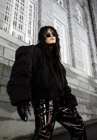 Sort oversized dunjakke med en tekstureret finish, kombineret med skinnende sorte vinylbukser. Tilbehør omfatter mørke solbriller og handsker.