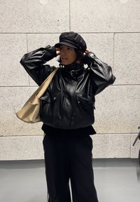 Mustast nahast jakk kõrge krae ja esitaskutega, koos musta nokamütsi ja beežika õlakotiga, taustal tekstureeritud sein.