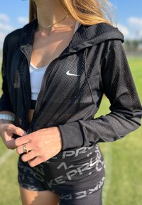 Nike crna mrežasta jakna s kapuljačom, s prednjim patentnim zatvaračem, u kombinaciji s crnim šortsevima koji prikazuju bijeli uzorak Nike logotipa.