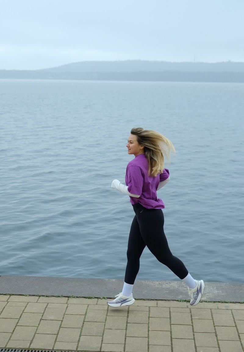 Jeune femme courant le long d'un sentier au bord du lac, portant une veste violette, un legging noir et des chaussures de course blanches par une journée nuageuse.