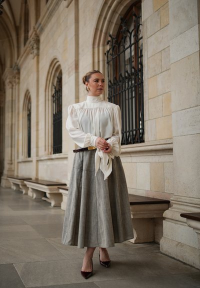 Mujer con blusa blanca y falda larga gris, de pie en un corredor arqueado de piedra con ventanas con rejas de hierro, mirando hacia un lado.