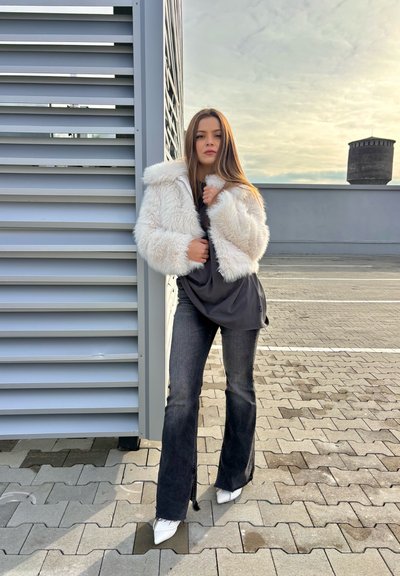 Chaqueta corta de piel sintética blanca, top gris, jeans oscuros acampanados y tacones blancos puntiagudos. La modelo está de pie contra una estructura gris en un pavimento con patrón.