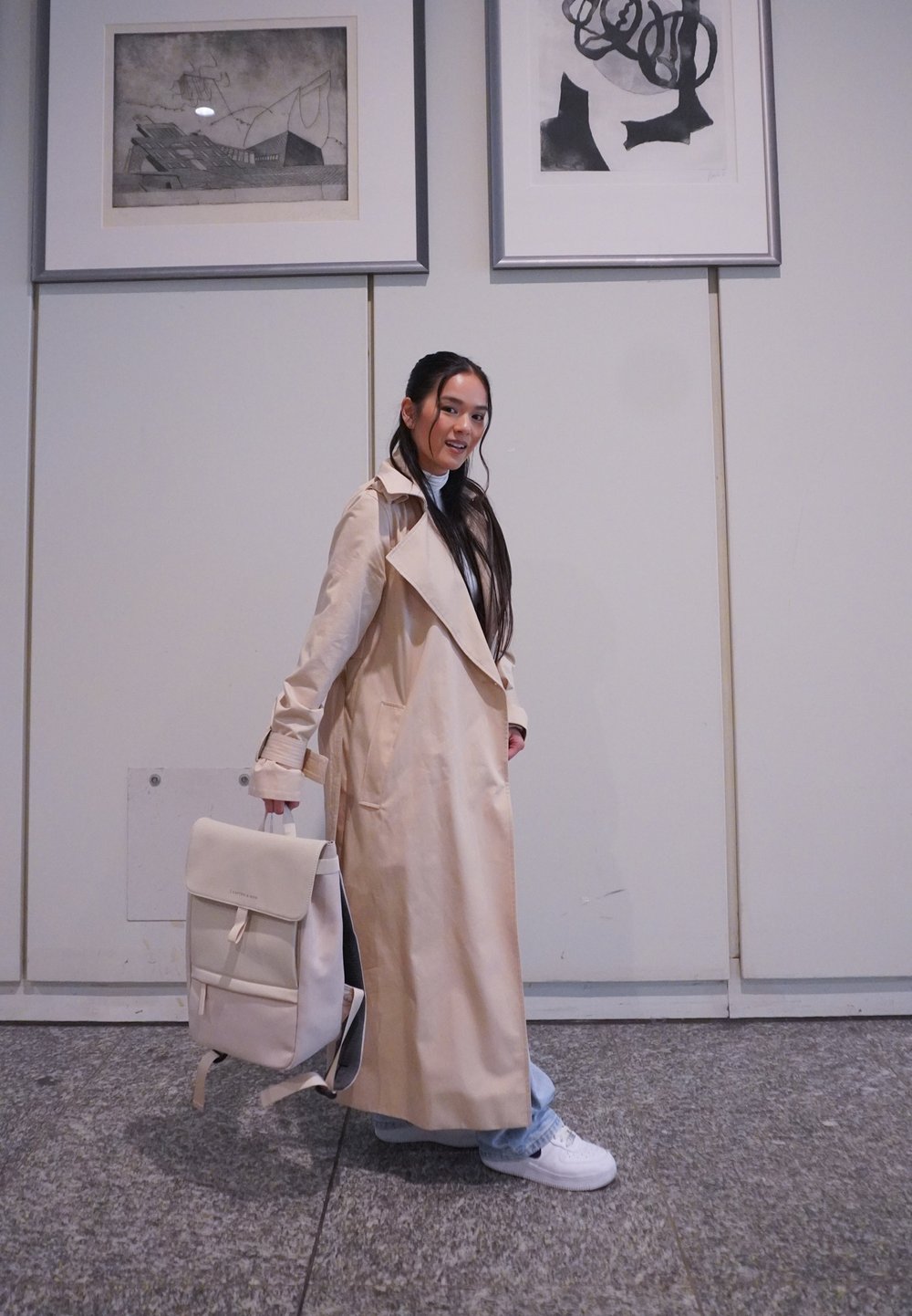 Gabardina beige con solapas anchas, mangas largas y bolsillos laterales. Sostiene una mochila de color claro con solapa y correas ajustables.