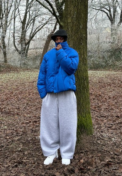 Chaqueta azul tipo puffer, diseño corto, patrón acolchado. Sudaderas gris de gran tamaño, zapatillas blancas, con un gorro negro y las manos cerca de la cara.