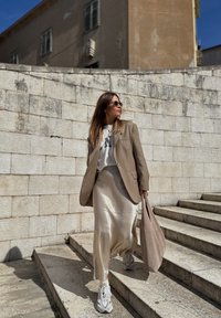 Blazer bege estruturado sobre uma t-shirt gráfica branca, saia de seda bege e ténis brancos. Bolsa tote castanha, fundo de parede de pedras multicoloridas.