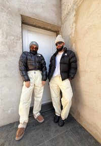 Δύο άτομα στέκονται δίπλα σε μια πόρτα, φοράνε μαύρες μπουφάν, ανοιχτό μπεζ αδιάβροχα παντελόνια και σκουφάκια. Ο ένας φοράει πορτοκαλί γυαλιά ηλίου, ενώ ο άλλος έχει ένα λευκό μπλουζάκι.