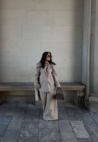 Beige oversized blazer met een sjaaldetail, gecombineerd met wijde broek in dezelfde stof. Donkere handtas, grote zonnebril en stenen achtergrond.