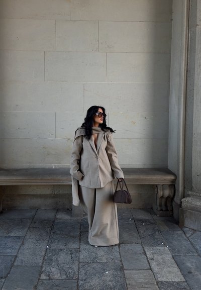 Blazer beige de gran tamaño con un detalle de bufanda, combinado con pantalones de pierna ancha en tejido a juego. Bolso oscuro, gafas de sol grandes y fondo de piedra.