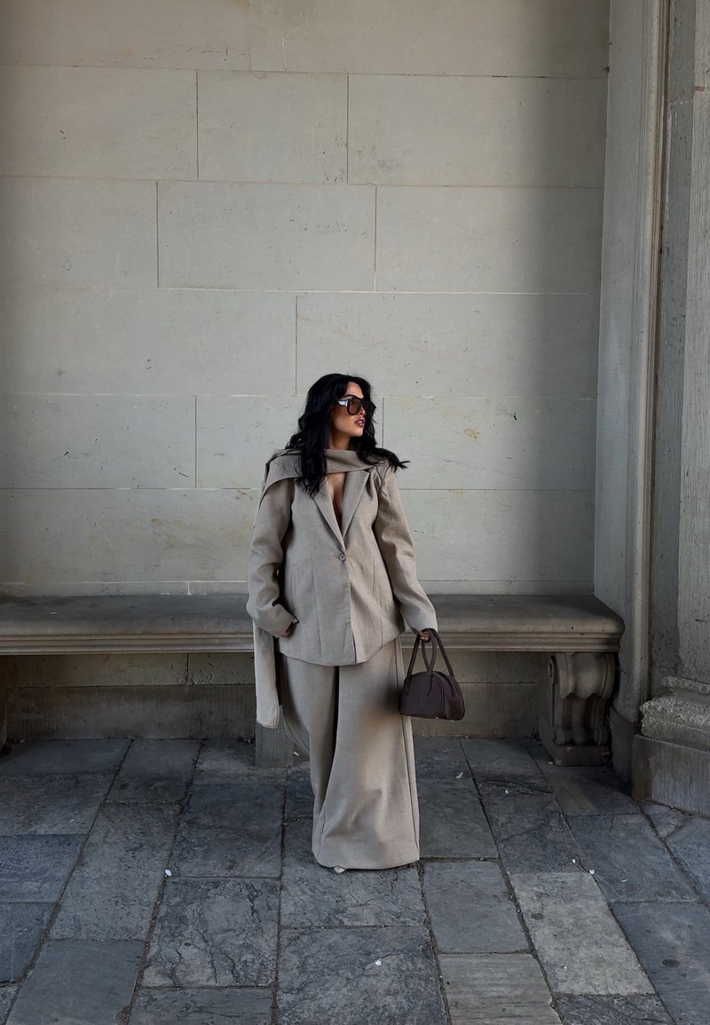 Beige Oversized-Blazer mit Schal-Detail, kombiniert mit weit geschnittenen Hosen aus passendem Stoff. Dunkle Handtasche, große Sonnenbrille und steiniger Hintergrund.
