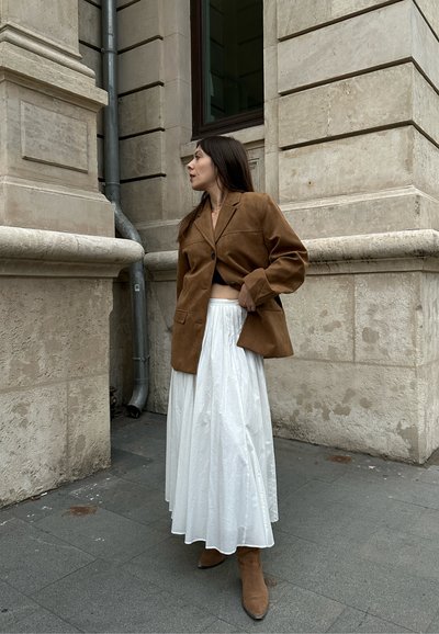 Blazer de ante marrón, falda blanca con pliegues y botines de ante color tostado. El blazer tiene un collar en pico y bolsillos frontales, mientras que la falda presenta un acabado texturizado.