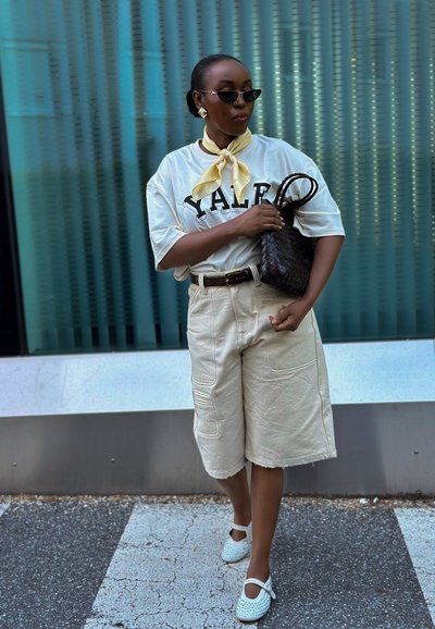 Camiseta oversized crema con estampado "Yale", pantalones cortos beige hasta la rodilla, zapatos bl blancos perforados, bolso negro de mimbre, bufanda de seda amarilla.