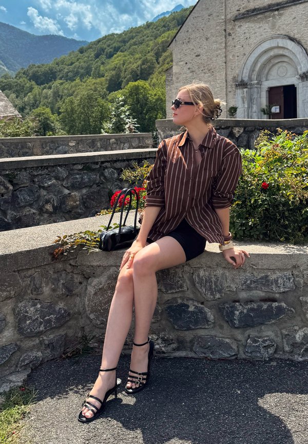Jeune femme portant des lunettes de soleil et une chemise rayée, assise sur un mur en pierre près de fleurs avec des montagnes et un vieux bâtiment en arrière-plan.