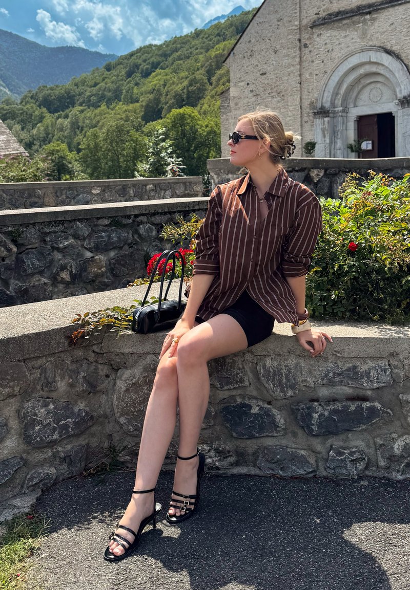 Jeune femme portant des lunettes de soleil et une chemise rayée, assise sur un mur en pierre près de fleurs avec des montagnes et un vieux bâtiment en arrière-plan.