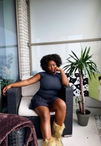 Silla de ratán negra con cojines blancos, una parte superior texturizada de color azul oscuro, falda oscura en capas y botas de piel de color beige. Planta en maceta y pared estampada en el fondo.