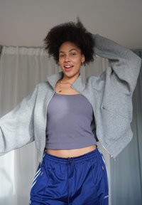 Chaqueta gris recortada con cremallera y mangas anchas sobre una camiseta sin mangas ajustada morada, combinada con pantalones cortos deportivos azules con franjas laterales blancas.