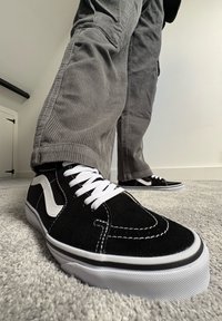 Schwarze Wildlederschuhe für Skateboarding mit weißen Schnürsenkeln und Nähten, ausgestattet mit einem weißen Seitenstreifen und einer schwarzen Gummisohle, auf einem grauen strukturierten Teppich.