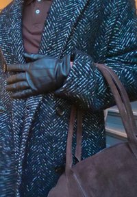 Czarny, teksturowany płaszcz w jodełkę, zestawiony ze stylowymi czarnymi skórzanymi rękawiczkami i ciemną zamszową torebką. Widoczne detale guzików.