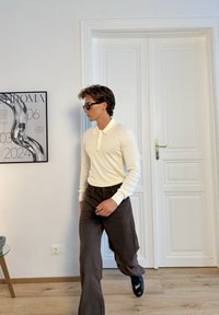 Biely polo s dlhým rukávom a golierom, kombinovaný s tmavohnedými širokými nohavicami a čiernymi topánkami. V pozadí je svetlá drevená podlaha a biele dvere.