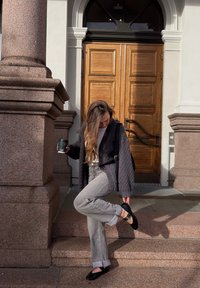 Svart strikket cardigan, hvit topp, grå bukser med rette ben og svarte flate sko. Står på trapp med en grønn kaffekopp, tre dører i bakgrunnen.