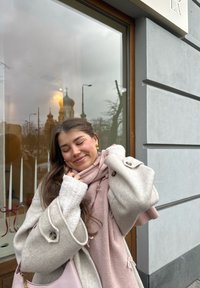 Jeune femme aux longs cheveux portant un manteau beige et une écharpe rose sourit les yeux fermés, debout devant une fenêtre reflétant le dôme d'une église et une rue.