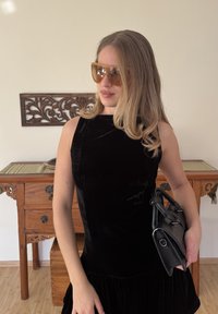 Robe sans manches en velours noir avec un bas évasé, associée à des lunettes de soleil surdimensionnées ambrées et un petit sac à main noir.
