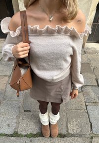 Sweater creme de malha off-shoulder com detalhe de babados, saia castanha com padrão xadrez, perneiras creme e botins castanhos, segurando uma mala castanha.