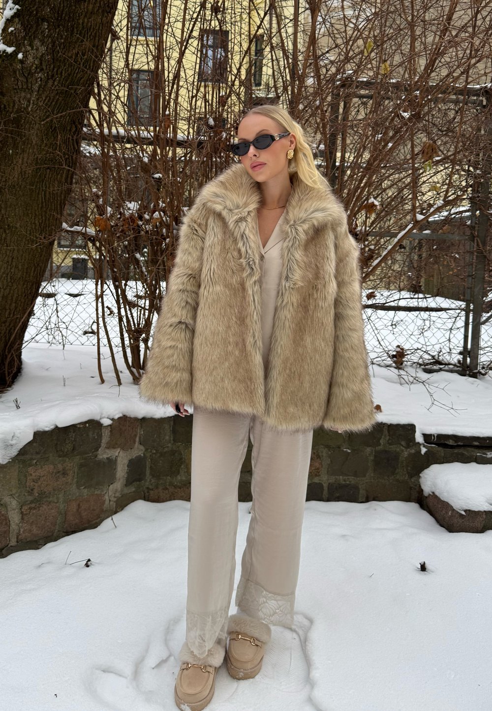 Femme portant un manteau en fourrure beige, un pantalon crème et des lunettes de soleil, debout sur la neige à côté d'un arbre et de buissons sans feuilles, avec un mur en pierre en arrière-plan.