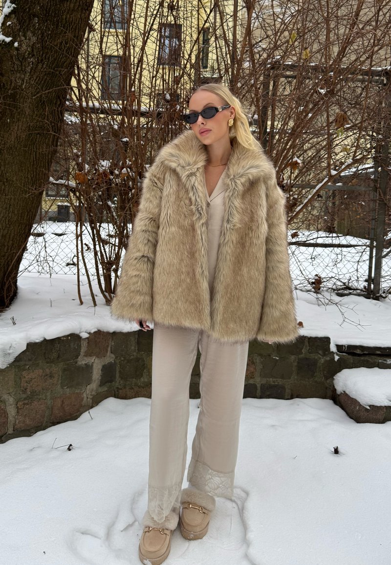 Femme portant un manteau en fourrure beige, un pantalon crème et des lunettes de soleil, debout sur la neige à côté d'un arbre et de buissons sans feuilles, avec un mur en pierre en arrière-plan.