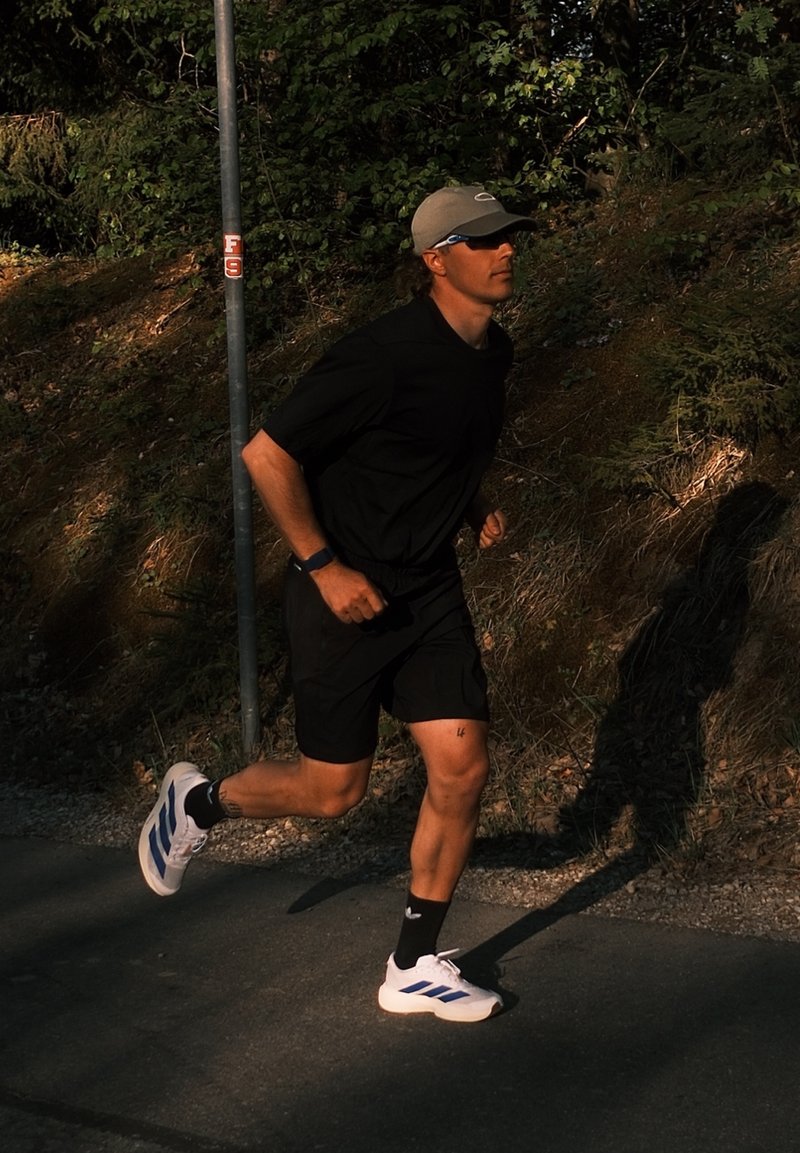 Homme portant des vêtements de course noirs et des baskets blanches, jogging sur un chemin ensoleillé à côté d'un feuillage vert en journée.