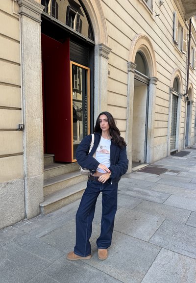 Pantalones de mezclilla de pierna ancha azul, chaqueta bomber negra, camiseta blanca con gráfico de animal, botas marrones y un bolso pequeño. De pie sobre un pavimento de piedra.