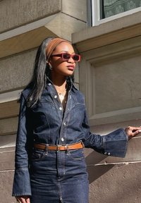 Abito camicia in denim con silhouette aderente, maniche lunghe a campana, bottoni a contrasto, messo in risalto da una cintura marrone e occhiali da sole rossi.