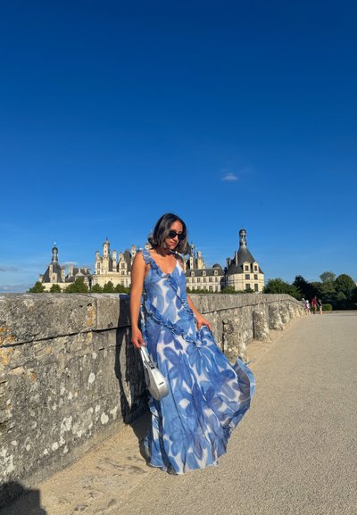 Vestido azul claro con patrones florales, bordes volados y una tela fluida. El fondo presenta un castillo histórico bajo un cielo azul despejado.