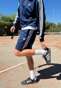 Tuta sportivo navy con strisce bianche, maniche corte e pantaloni corti. Sneakers nere con strisce bianche. Ambiente luminoso di un campo da tennis all'aperto.