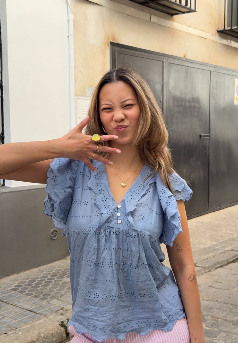 Femme en blouse bleue à œillets et pantalon rayé rose montrant la main avec des bagues tout en faisant une expression facétieuse sur un trottoir en ville.