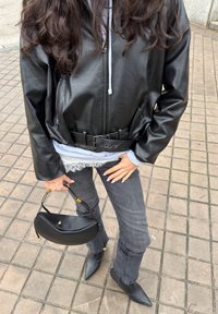 Personne portant une veste en cuir noire, un jean gris, des bottes noires à bout pointu, tenant un sac à main noir, debout sur un pavé extérieur carrelé.