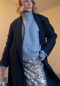Femme aux cheveux blonds courts portant un manteau noir, un pull gris zippé et une jupe en sequins argentés, tenant un sac à main en sequins assorti.